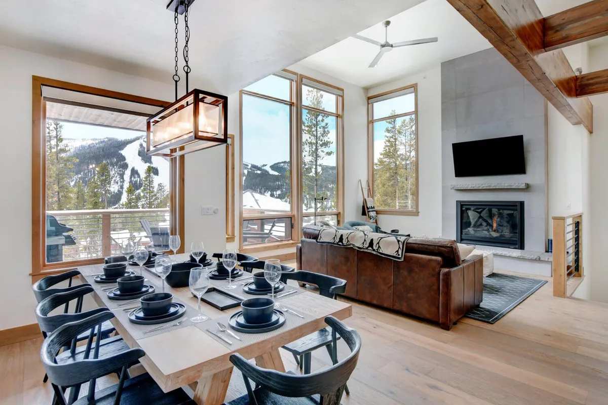 A contemporary dining area set for a meal, featuring a large table surrounded by comfortable chairs. Expansive windows offer a stunning view of the mountains, and a warm-toned couch and fireplace add to the inviting atmosphere. The smart TV positioned above the fireplace is also visible, linking the living and dining zones.