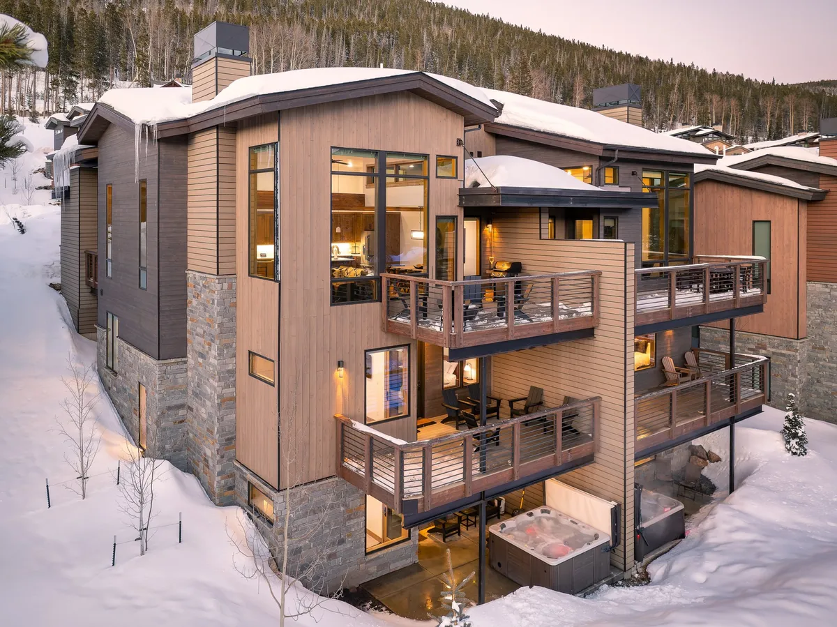 The image showcases a luxurious mountain villa in Winter Park, Colorado, with inviting wooden balconies, expansive windows, and a private hot tub bubbling on the deck. Snow blankets the roof and surrounding landscape, while the balcony is set up for outdoor dining and offers panoramic winter views—a cozy retreat against the stunning alpine backdrop.