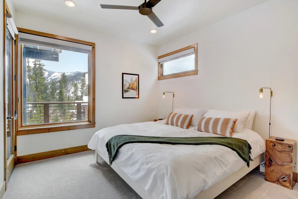 A peaceful bedroom with a large bed dressed in crisp white bedding, a wooden nightstand, a window showcasing beautiful mountain views, and tasteful decorative artwork.
