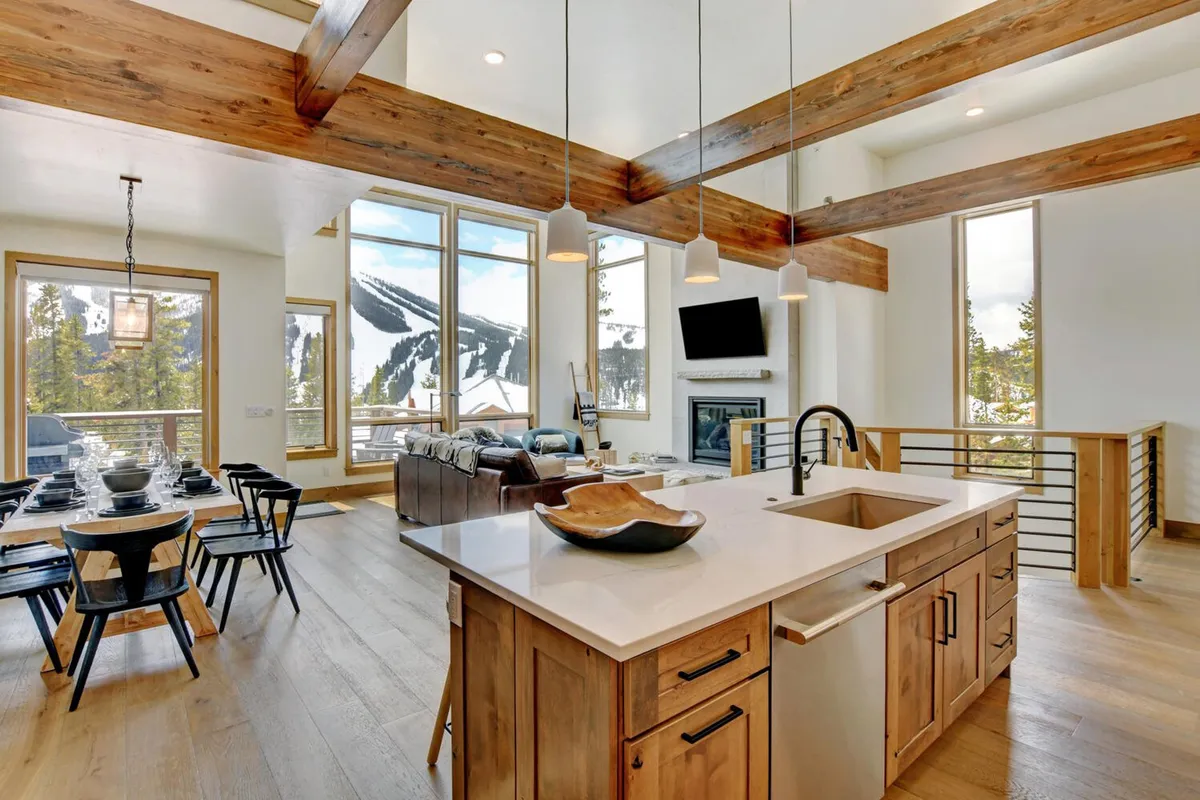 A comfortable kitchen and dining space featuring a kitchen island, a dining table set for a meal, large windows revealing mountain views, and exposed wood beams. The setup exudes a warm, inviting mood ideal for social gatherings.