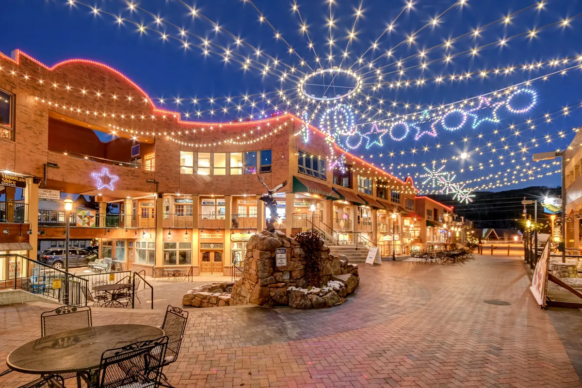 A vibrant downtown streetscape illuminated by twinkling lights and decorated with stars and snowflakes. Warmly lit storefronts line a brick pathway, inviting guests for evening strolls, outdoor dining, boutique shopping, and festive celebrations. The scene feels magical and inviting.
