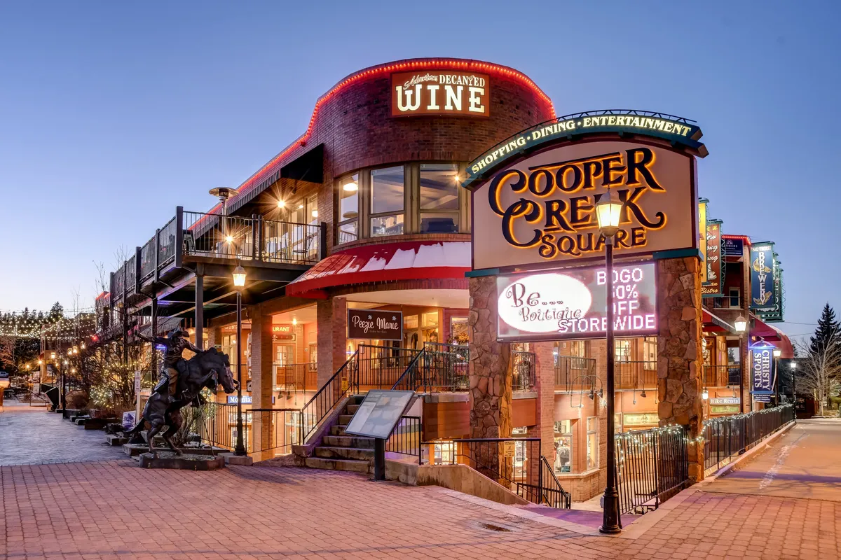 A lively exterior shot of Cooper Creek Square features signage, twinkling decorative lighting, and a staircase leading to a wine shop entrance. The scene evokes a sense of vibrant nightlife, seasonal festivals, and the opportunity for friends to explore local dining, shopping, and entertainment together.