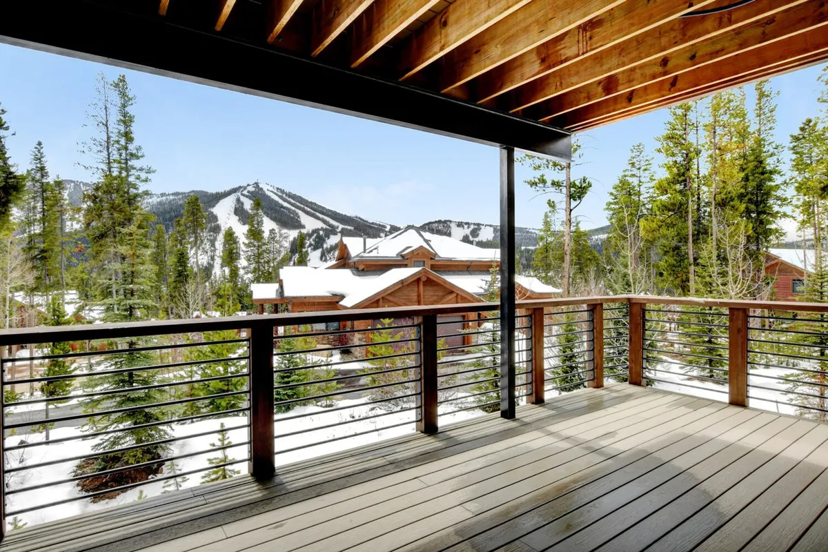 A modern deck overlooks a stunning mountain vista, with snow-covered trees providing a serene backdrop. The deck's railing frames the view, while guests relax, enjoying morning coffee or watching winter activities in the distance. The atmosphere is peaceful, inviting both quiet moments and shared gatherings.