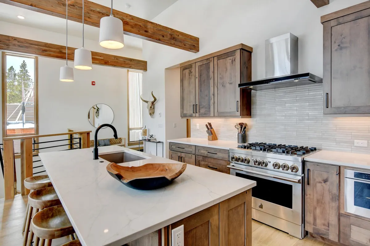 A warm, inviting kitchen with a large island at its center, surrounded by wooden beams and featuring stainless steel appliances. The island is topped with a decorative bowl and framed by bar seating, making it the heart of the space for cooking and conversation.