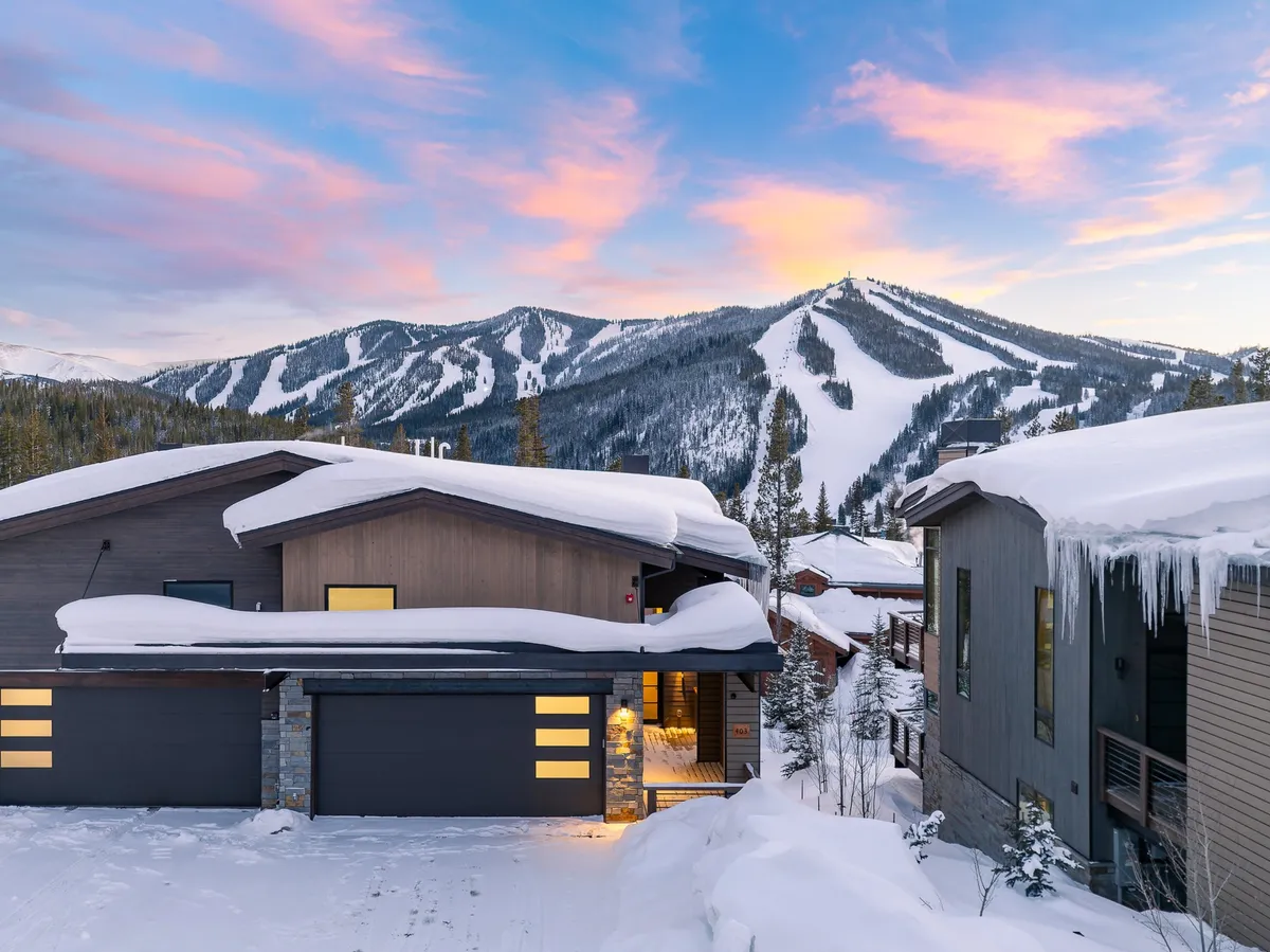 A cozy exterior winter scene featuring snow-covered rooftops set against a mountain backdrop. The warmly lit entrance hints at comfort inside, while people can be seen skiing, snowboarding, and relaxing by the fireplace, all beneath a beautiful sunset glow.