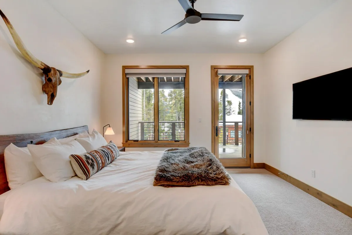 A tranquil bedroom featuring a king-sized bed with decorative pillows, a mounted animal head as wall decor, large windows with a view of the surrounding landscape, and a sliding glass door opening onto a private balcony.