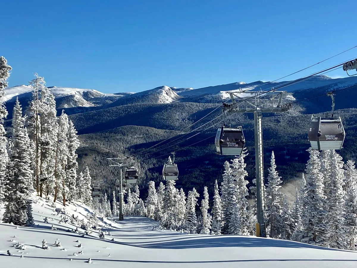 A dynamic winter scene featuring ski lift gondolas gliding above snow-covered trees, with rugged mountain peaks rising in the background. The landscape is blanketed in fresh snow, creating a sense of exhilaration and anticipation for on-mountain adventure.