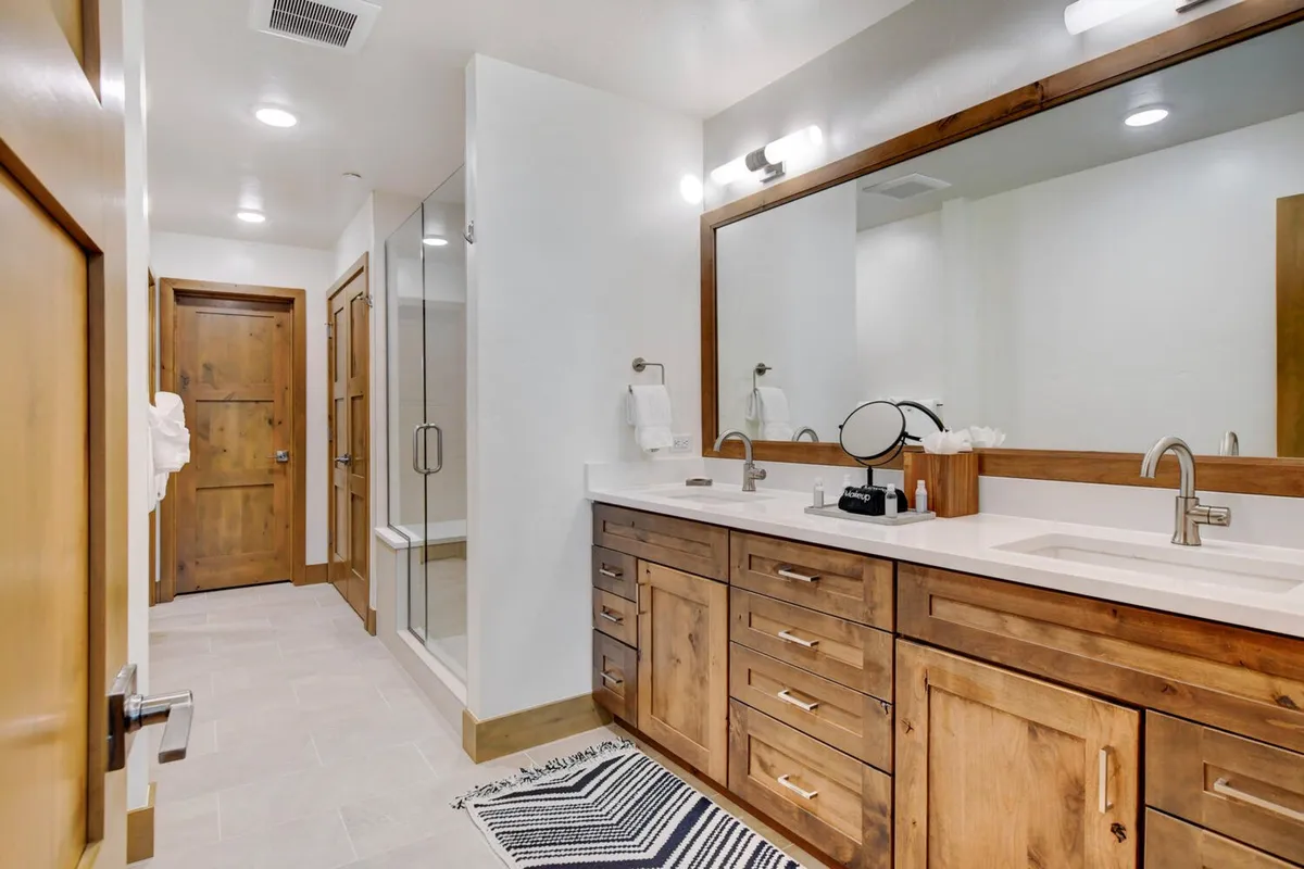 A spacious bathroom with a double sink vanity, large mirror, wooden cabinetry, a glass shower enclosure, and a soft textured rug, exuding an inviting and serene atmosphere.