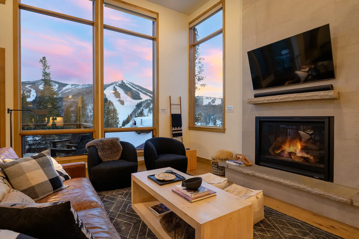 A spacious living area featuring large windows that perfectly frame the snow-capped Rocky Mountains, a plush seating area centered around a glowing gas fireplace, and soft lighting that enhances the warm, inviting mood. The overall ambiance is both cozy and grand, with the outdoors visually connected to the indoors.